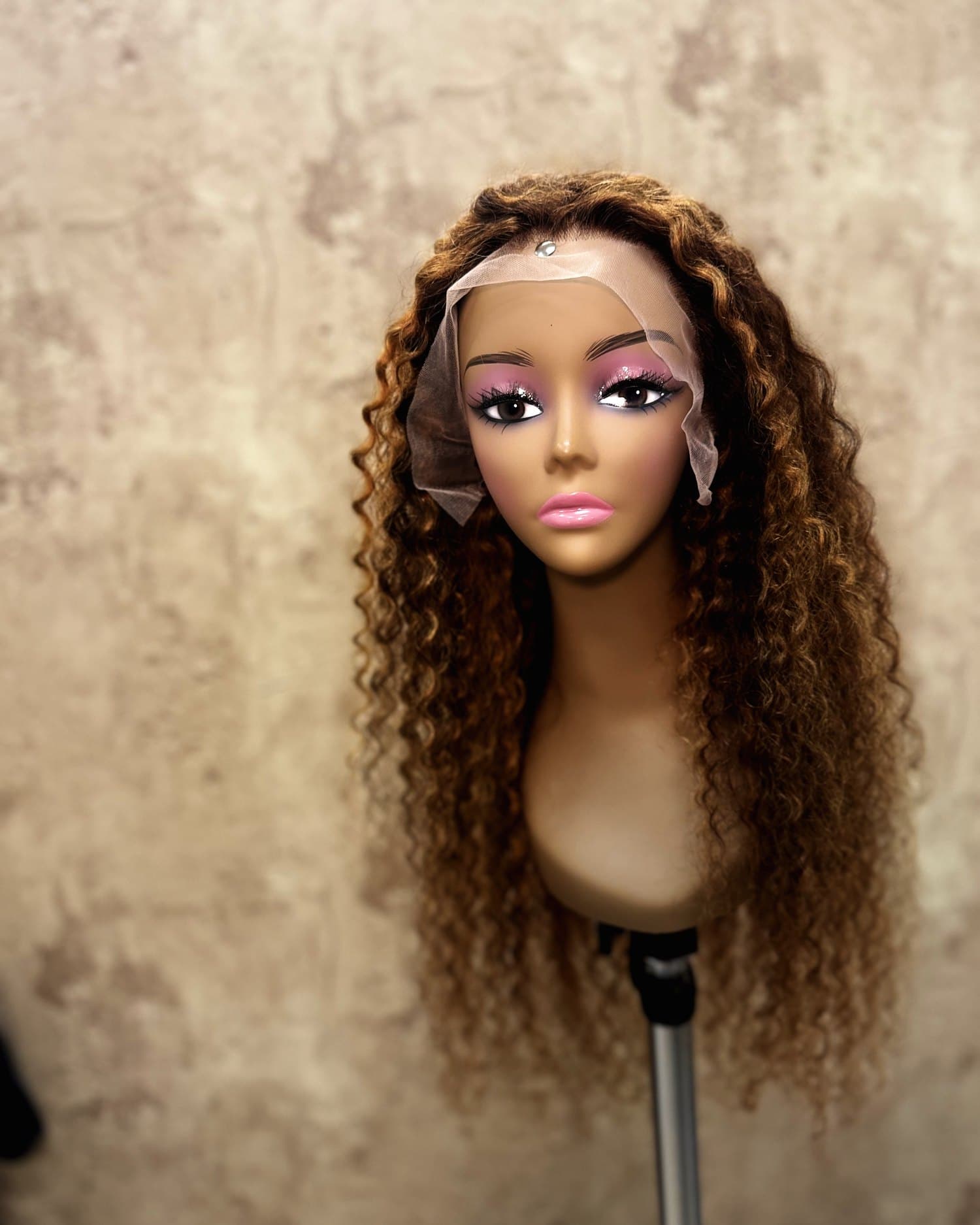 Honey-blonde curly lace unit on a stand, lace laid over the forehead.