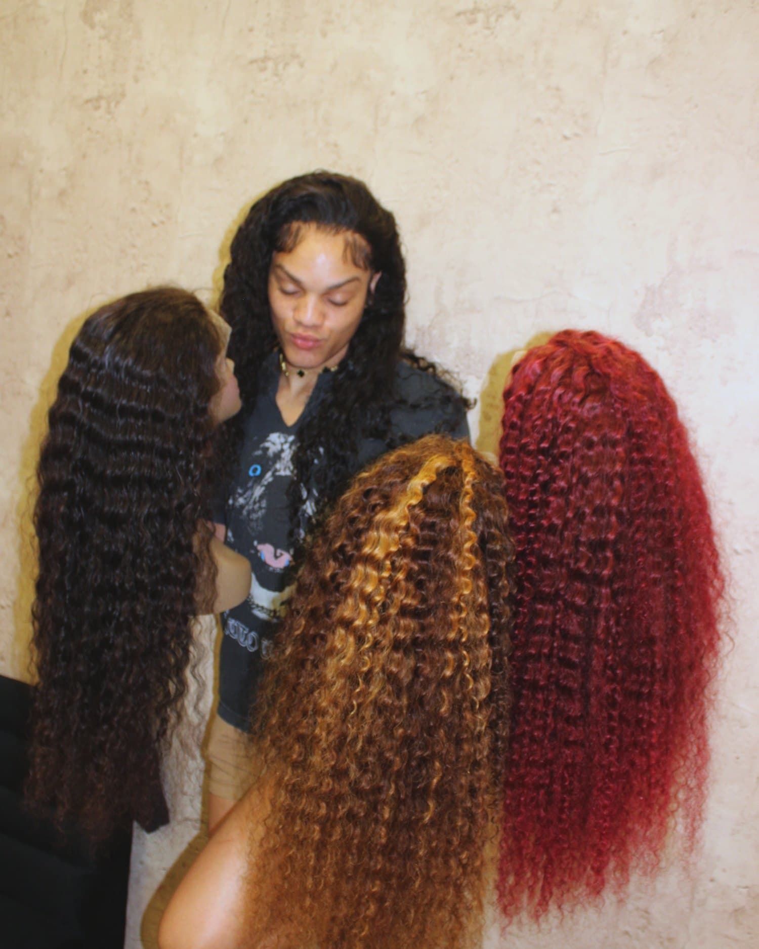 Quality holding three long units — black deep wave, honey curly and red — in her workroom.