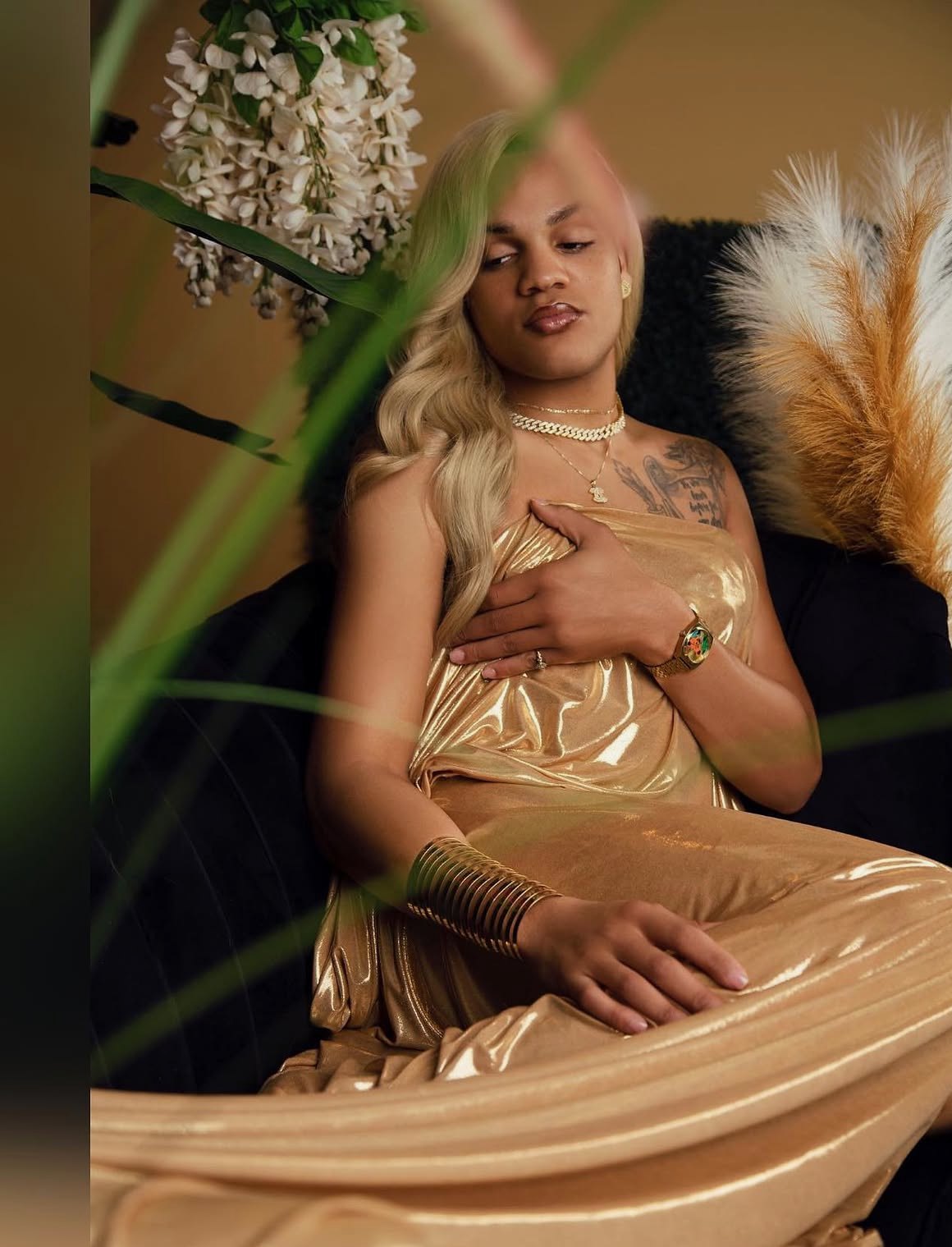 Quality, the stylist, in a gold gown with long blonde waves, reclining in a black chair